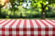© oksa_studio - Red and white checkered tablecloth on an empty table outdoors, ready for a summer picnic. Blurry green nature background with copy space