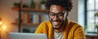 © miss irine - Man with glasses smiles while looking at laptop. He wears yellow sweater, sits at table. Person works remotely from home office. Technology use for communication and business.