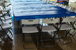© Semiglass - A long rectangular table covered with a bright blue disposable plastic tablecloth is encircled by white metal-frame folding chairs on a glossy tiled floor in a spacious indoor event area.