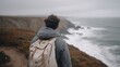 © CYBERPINK - Young man standing on a cliff overlooking the ocean. he is wearing a grey hoodie and a beige backpack. the man is facing away from the camera, with his back towards the horizon.