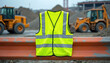 © Vadym - Bright yellow safety vest with reflective stripes hangs on orange traffic barrier. Construction site with heavy machinery, piles of dirt, buildings visible in background. Vest serves as personal