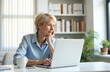 © Pete - Mature woman using laptop computer at desk, thinking while typing. She works remotely from home office, looking thoughtful and engaged in her task. White background with bookshelves.
