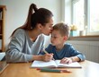 © Pete - Mother kisses forehead of young son doing homework at table. Child learns indoors with pen and book. Family shows love and support during study time at home.