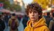 © Pete - Young man with curly brown hair wears yellow hoodie. He stands in a busy city street full of people. Natural light illuminates his thoughtful face and casual style.