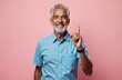 © Viktor - Smiling senior man raises index finger, signifies idea or solution. Experienced mature guy with gray hair and beard, light blue shirt, isolated on pink.