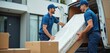 © Viktor - Two male movers in blue uniform carry wrapped mattress from house to van. They smile and work together, loading furniture for delivery. Boxes are nearby.