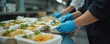 © Viktor - Kitchen workers in blue gloves prepare meals in takeaway containers. Chefs assemble food orders for delivery service. Employees pack lunch boxes in commercial kitchen.