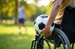 © Viktor - Person in wheelchair holds soccer ball, playing with friend in park. Active disabled man enjoys sport recreation with buddy outdoors on green grass field.