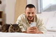 © Pixel-Shot - Handsome young man with mobile phone and cute cat lying on bed in bedroom