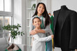 © Pixel-Shot - Happy Asian mother and her daughter taking measurements of black jacket on mannequin in sewing studio