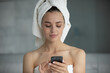 © fizkes - Morning routine and digital habits. Head shot portrait relaxed young female with towel around body turban on wet hair text messages on phone surf internet after self care hygienic shower procedures