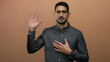 © Krakenimages.com - Young hispanic man in glasses with one hand on chest and the other raised against a brown wall, showcasing a gesture of honesty in a studio portrait.