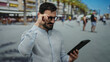 © Krakenimages.com - Man with beard using tablet outdoors on sunny street, adjusting glasses, showcasing hispanic ethnicity and business casual attire in lively urban environment.