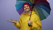 © Krakenimages.com - Young man in yellow raincoat holds colorful umbrella against isolated purple background, extending palm with a confident, handsome expression and charming demeanor.