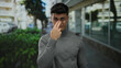 © Krakenimages.com - Young man makes a 'watching you' gesture on a city street, wearing glasses and a gray sweater, with blurred urban scene in background.