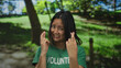 © Krakenimages.com - Young chinese volunteer woman with crossed fingers in park setting, wearing green t shirt and smiling, surrounded by lush outdoor scenery.