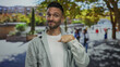 © Krakenimages.com - Young hispanic man pointing to himself confidently on a vibrant urban street with blurred people and green trees in the background.