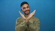 © Krakenimages.com - Young man in a green sweater crosses his hands in an x gesture against a solid blue background indicating rejection with a confident expression.
