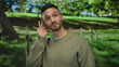 © Krakenimages.com - Young hispanic man in a green park outdoors showing a listening gesture with a curious expression and thumbs up, surrounded by vibrant nature under a clear blue sky.