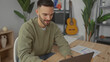 © Krakenimages.com - Hispanic man working on a laptop in a cozy living room setting with a guitar and houseplants nearby, embodying a relaxed home office atmosphere during daytime.