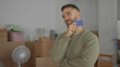© Krakenimages.com - Young man indoors in new home holding a blue credit card surrounded by moving boxes looking thoughtful in a casual environment with cardboard and minimal furniture around him