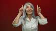 © Krakenimages.com - Senior woman with grey hair in white shirt points upwards against red background indicating excitement or focus and isolated in studio setting.