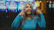 © Krakenimages.com - Senior woman with grey hair smiles while pointing to her cheeks inside a vibrant casino with colorful slot machines in the background, creating a cheerful indoor gaming atmosphere