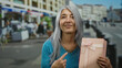 © Krakenimages.com - Woman with grey hair points at pink gift box while standing on a busy street, highlighting her joyful expression and the vibrant urban environment around her.