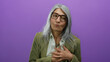 © Krakenimages.com - Senior woman with grey hair and glasses clutching chest in discomfort against isolated purple background, conveying health concern and emotion.