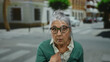 © Krakenimages.com - Senior woman with grey hair and glasses stands outdoors on a street in a city setting, expressing surprise or confusion while pointing at herself.