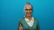 © Krakenimages.com - Senior woman with grey hair and glasses smiles giving thumbs up against blue background suggesting positivity and approval.