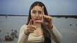 © Krakenimages.com - Woman holds a loser sign with her hand toward the camera on a windy beach with scattered rocks under an overcast sky at low tide; mockery.