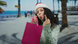 © Krakenimages.com - Woman wearing santa hat holding pink gift bag on promenade near beach showcasing playful gesture against sea backdrop in sunny weather