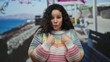 © Krakenimages.com - Young hispanic woman on a promenade by the sea, wearing a colorful sweater and pointing at herself with a surprised expression against a scenic beach backdrop.