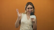 © Krakenimages.com - Woman holding microphone and waving hand in orange studio with plain backdrop, facing camera in mid shot; cheerful greeting.