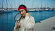 © Krakenimages.com - Woman with tennis racket standing outdoors at a port with boats, wearing a red headband and white sweatshirt, expressing confident smile on a sunny day.