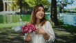 © Krakenimages.com - Woman holding flowers in a park with vibrant green trees, wearing a white shirt with a tattoo on her arm, expressing gratitude and happiness under sunny weather.