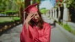© Krakenimages.com - Young woman in pink graduation gown appears worried in a sunny park setting, showcasing concern and contemplation on her face, with a blurred path in the background.