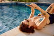 © we.bond.creations - Woman lying poolside by a blue swimming pool, relaxing and checking her phone during a tropical summer holiday