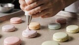 Close-up of hands piping cream onto colorful macarons on a baking sheet.