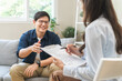 © KMPZZZ - Psychology, depression. smile asian young male, man patient mental sitting on couch, explained symptom with psychologist, psychiatrist while doctor woman taking notes at clinic. Encouraging, therapy.