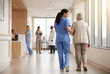 © simohamed - A nurse in blue scrubs gently assists an elderly female patient walking down a bright hospital corridor, with other medical staff and patients in the background.