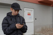 © Viktar - Teenager checking messages near garage door, Teenage boy wearing jacket and cap reviews messages outside, Young white male reviews messages on his phone near garage with city surroundings