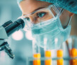 © Balian Leming - Close-up of a female lab technician wearing safety gear and a surgical mask using a microscope with test tubes visible in foreground.