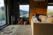 © Home-stock - Lady relaxing on sofa with book and enjoying mountain views from large window, cozy room with firewood stack in the corner, free space