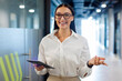 © Home-stock - Joyful businesswoman standing in office hallway, woman economist accountant looks joyfully at camera and holding digital tablet