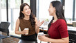 © NAMPIX - Two cheerful Asian businesswomen enjoying a coffee break and chatting in a bright modern office workspace.