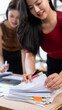 © NAMPIX - Two professional Asian women collaborating and reviewing documents on a desk in a modern office workspace.