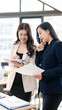© NAMPIX - Two Asian businesswomen analyzing market data on a digital tablet and documents in a modern office. Concept of professional teamwork and leadership.