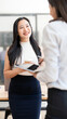 © NAMPIX - Smiling Asian businesswoman holding a coffee cup and tablet while chatting with a colleague in a bright office. Concept of positive office lifestyle.